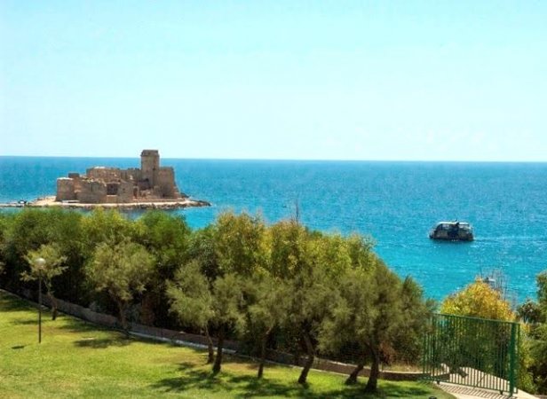 Un castello su un'isola vicino al mare limpido, con alberi verdi in primo piano.