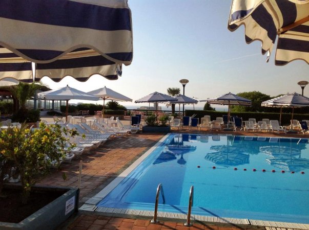 Piscina con sedie a sdraio sotto ombrelloni a righe blu e bianche, e un cielo limpido.