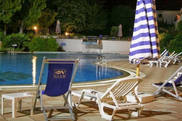 Piscina illuminata di notte, circondata da sedie a sdraio e ombrelloni a righe bianche e blu.