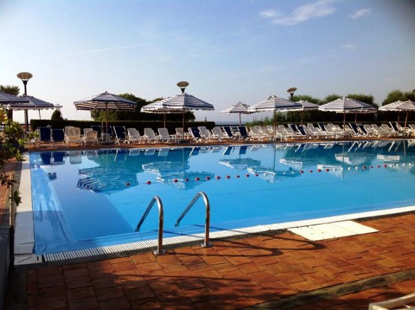 Piscina rettangolare con lettini e ombrelloni a righe bianche e blu. Cielo sereno sopra.