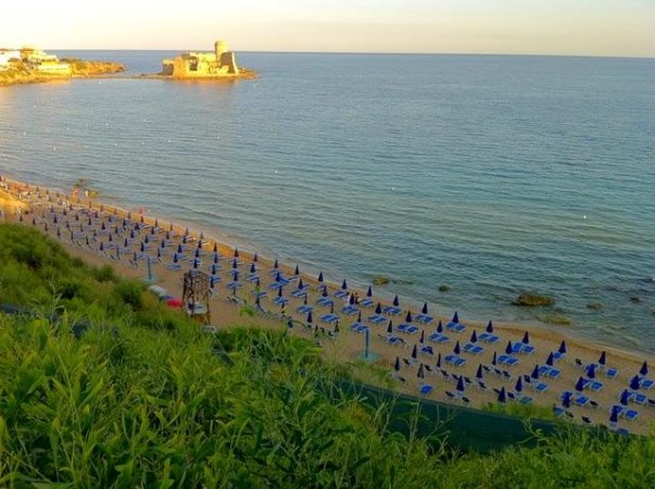 Spiaggia con file di ombrelloni blu lungo la costa e un castello in lontananza.