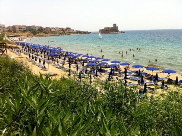 Spiaggia affollata con ombrelloni blu e un castello visibile in lontananza nel mare.