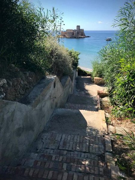 Sentiero scende verso la spiaggia, con un castello su un'isola in lontananza.