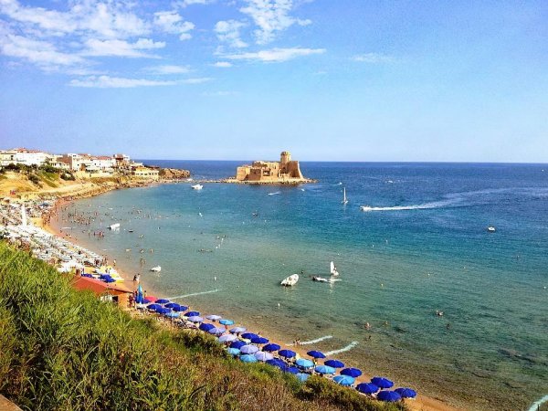 Spiaggia affollata con ombrelloni blu, castello in lontananza.