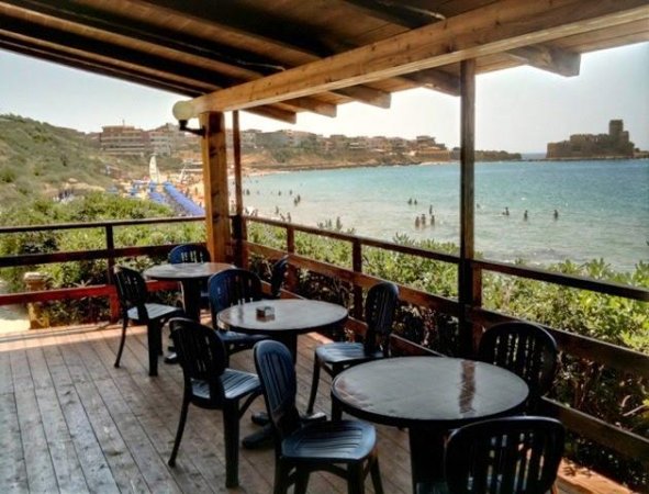 Terrazza con tavoli e sedie, vista mare e bagnanti in acqua.