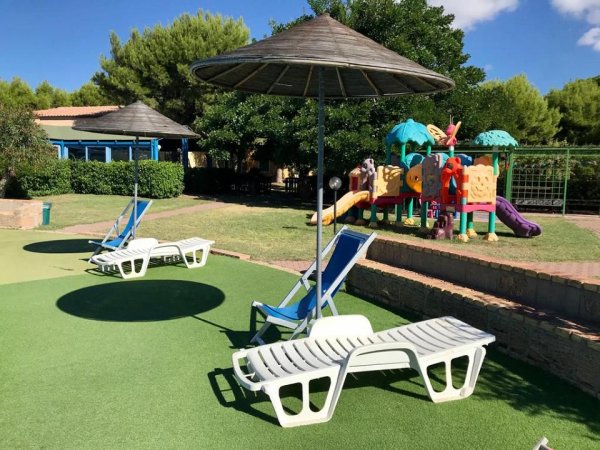 Lettini bianchi e ombrelloni su prato verde, con area giochi per bambini sullo sfondo.