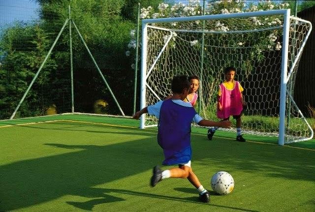 Bambini che giocano a calcio su un campo verde con porta e reti protettive.