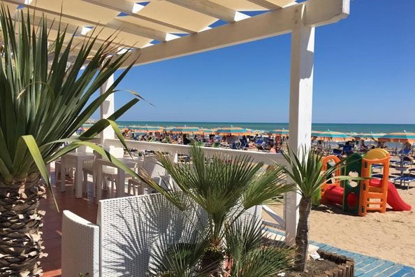 Veduta di una spiaggia affollata con palme e tavoli all'ombra.