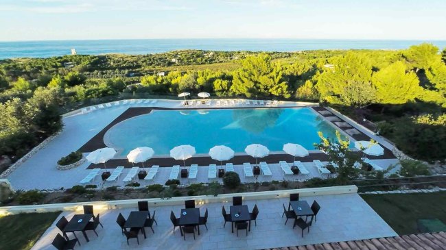 Vista di una piscina con ombrelloni, circondata da verde e vista mare.