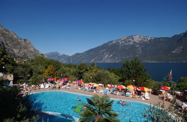 Piscina con persone e ombrelloni colorati, circondata da montagne e lago.