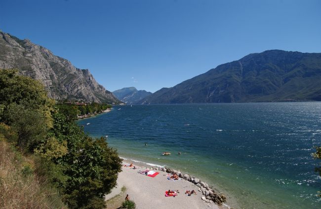 Spiaggia di ciottoli con persone, immersa tra montagne e lago.