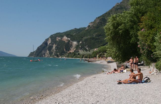 Spiaggia di ciottoli con bagnanti e montagne, accanto ad alberi verdi.