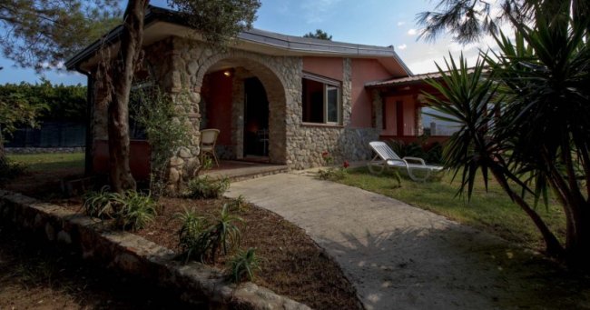 Casetta in pietra con portico, sdraio e giardino curato con piante.