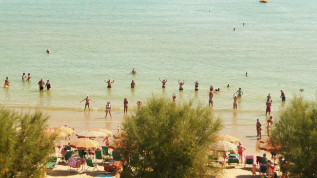 Persone in acqua poco profonda su una spiaggia con ombrelloni colorati e vegetazione.