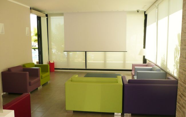 Sala con sedie colorate, grande schermo bianco e finestre luminose.