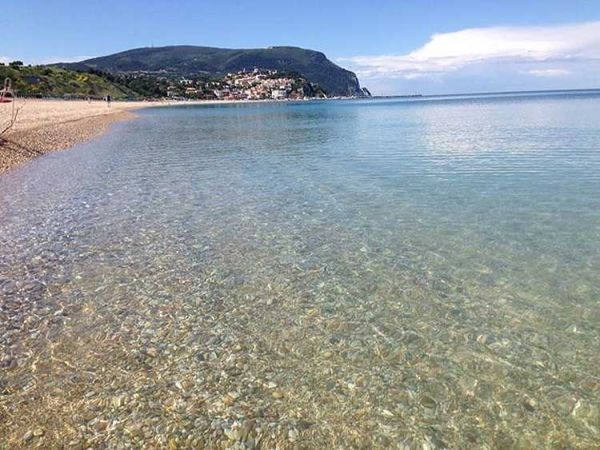 Spiaggia di ciottoli con mare cristallino e colline sullo sfondo.