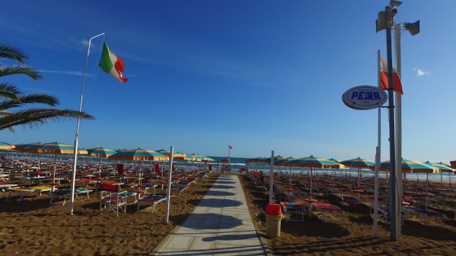 Spiaggia con lettini e ombrelloni colorati, bandiera italiana al vento.