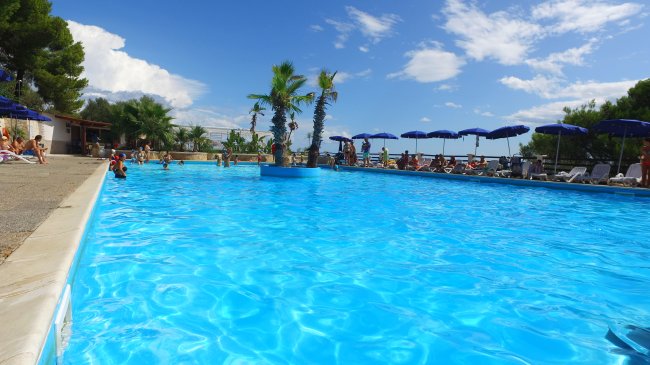 Piscina con palme e ombrelloni blu sotto un cielo soleggiato.