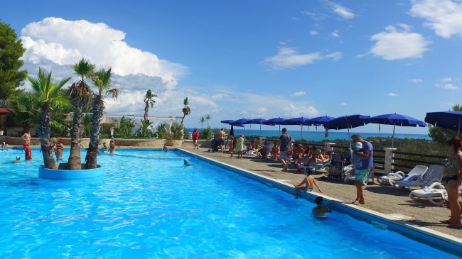 Bordo piscina con ombrelloni blu e persone che si rilassano al sole.