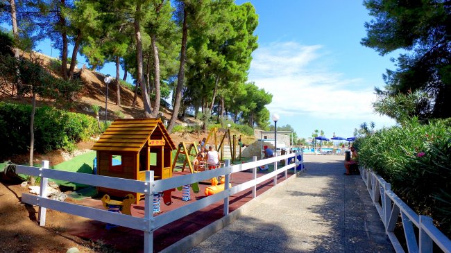 Area giochi per bambini con casetta e altalene, circondata da alberi.
