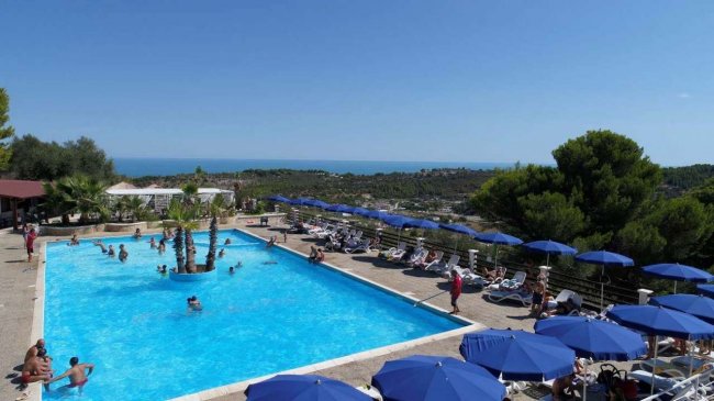 Una grande piscina all'aperto con ombrelloni blu, circondata da lettini, vista mare.