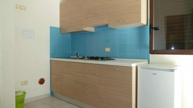 Cucina con mobili in legno chiaro, piastrelle blu e un piccolo frigorifero bianco.