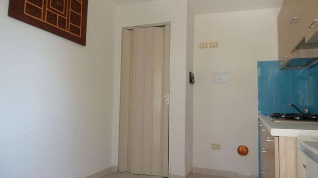 Angolo di una stanza con porta pieghevole beige, quadro appeso e parte della cucina.