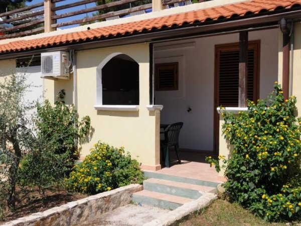 Piccola casa con tetto in tegole, veranda, e cespugli di fiori gialli.
