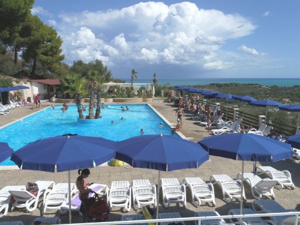 Piscina all'aperto con lettini e ombrelloni blu, vista sul mare e cielo nuvoloso.