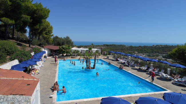 Piscina all'aperto con ombrelloni blu, vista sul mare e paesaggio collinare.