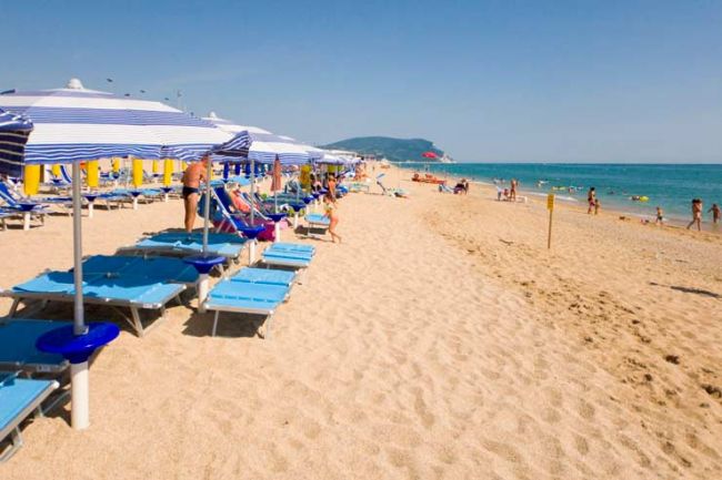 Lettini e ombrelloni blu disposti lungo la spiaggia, pochi bagnanti in acqua.