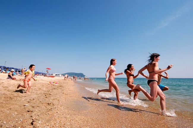 Ragazzi corrono verso il mare lungo una spiaggia sabbiosa.
