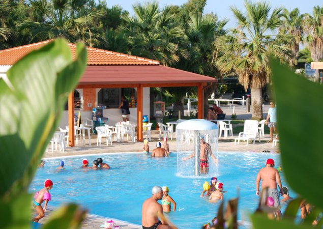 Piscina con spruzzi d'acqua e persone che nuotano, circondata da palme.