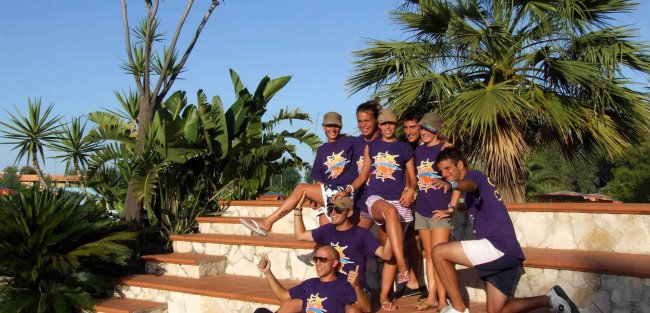 Gruppo sorridente con t-shirt viola seduto su gradini tra piante tropicali.