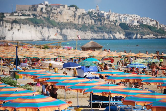 Spiaggia affollata di ombrelloni colorati, città sullo sfondo.