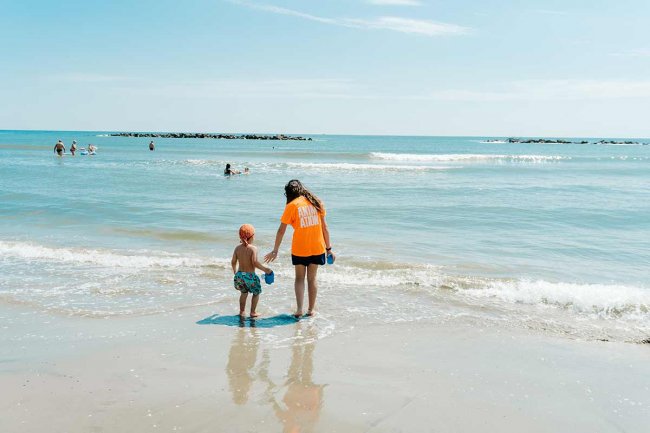 Una donna e un bambino camminano verso il mare, entrambi con secchielli colorati in mano.