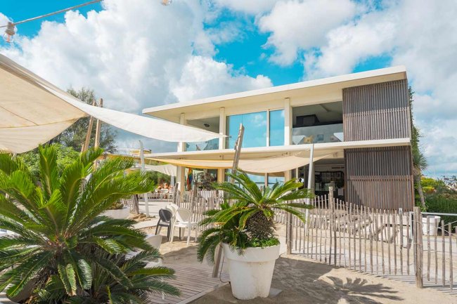 Edificio moderno con terrazza, sedie bianche e piante tropicali, cielo blu con nuvole.