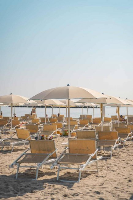 Una spiaggia con ombrelloni beige e lettini di legno su sabbia sotto un cielo sereno.