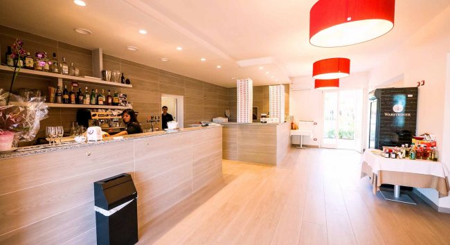 Un bar moderno con bancone in legno chiaro, luci rosse e scaffali con bottiglie dietro.