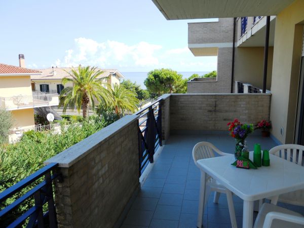 Terrazza con tavolo bianco e sedie, vista mare e vegetazione.
