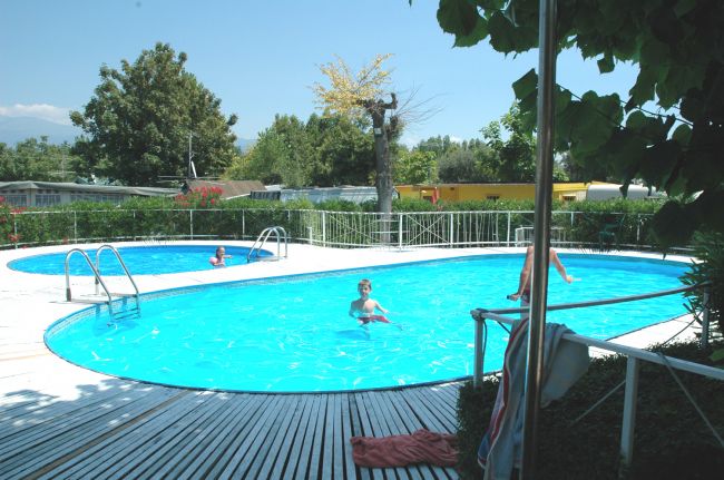 Piscina con deck bianco e persone che nuotano, circondata da alberi e siepi.