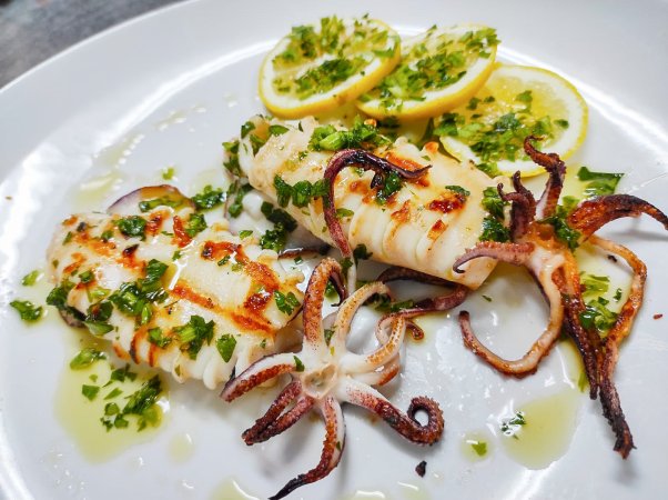 Calamari grigliati con prezzemolo e fette di limone su un piatto bianco.