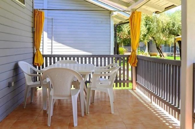 Una veranda esterna con tavolo bianco e sedie in plastica. Tende gialle e recinzione grigia.
