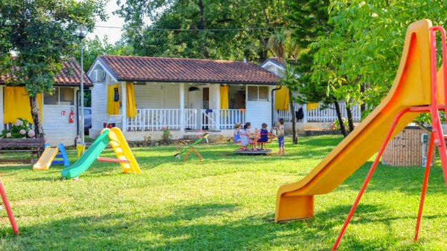 Parco giochi con scivolo arancione e casette bianche sullo sfondo.
