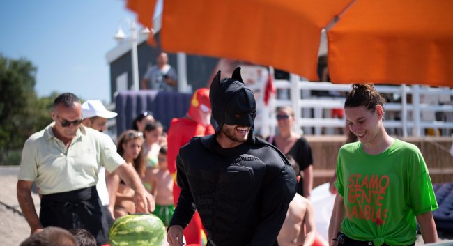 Persone in spiaggia con un uomo in costume da supereroe e ombrelloni arancioni.