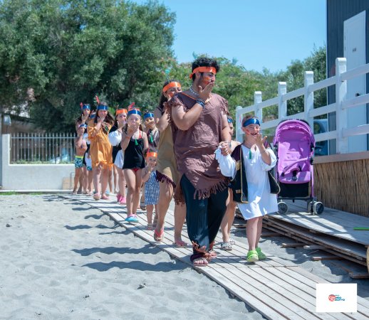 Un gruppo di bambini in fila con copricapi colorati cammina su una passerella di legno.