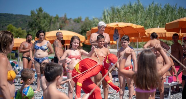 Bambini giocano con corde su una spiaggia, mentre gli adulti osservano sotto ombrelloni arancioni.
