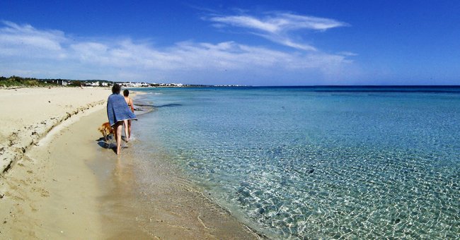 Due persone camminano su una spiaggia sabbiosa vicino al mare trasparente sotto un cielo azzurro.