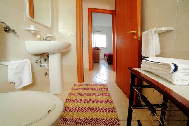 Bagno con lavabo bianco, asciugamani e tappeto colorato, vista su camera da letto.
