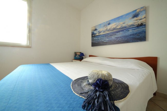 Letto matrimoniale con copriletto blu, un quadro del mare e un cappello di paglia posato sul letto.